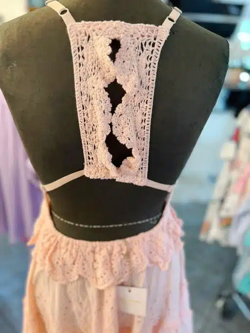 Eine Rückansicht einer schwarzen Schneiderpuppe mit einem rosa Cloe-Boho-Kleid kurz aus Spitze. Das Kleid weist aufwendige Häkelmuster auf der Rückseite und dekorative Spitzendetails um die Taille auf. Der Hintergrund zeigt verschwommene, mehrfarbige Kleidungsstücke.