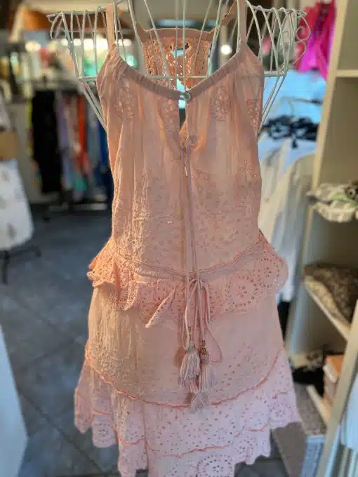 Ein rosa, ärmelloses Cloe Boho Kleid kurz mit zarten Spitzendetails und einer Quaste zum Binden auf der Rückseite wird auf einer Metallpuppe in Cloes Boutique ausgestellt. Im Ladenhintergrund sind Kleiderständer und Regale zu sehen.