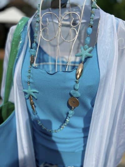 Eine Nahaufnahme einer Schaufensterpuppe in einem hellblauen Kleid, geschmückt mit verschiedenen Halsketten mit goldenen Anhängern wie Seesternen und Perlen. Der Fokus liegt auf dem