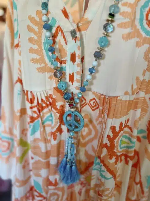 Eine Nahaufnahme eines bunt gemusterten Kleides mit einer „Summer Peace Boho“-Kette mit blauen Perlen und einem großen blauen Anhänger mit einer Quaste, präsentiert auf einer Schaufensterpuppe.