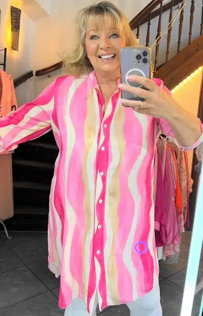 Eine Person macht ein Selfie im Spiegel, lächelt, trägt ein rosa-weiß gestreiftes Cara Tunika-Kleid und hält ein Smartphone in der Hand.