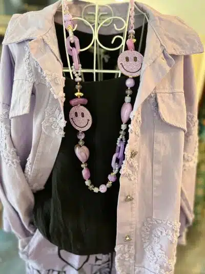 Eine lavendelfarbene Bluse mit Spitzenbesatz und einer Jeansjacke von Heard hängt auf einem weißen Kleiderbügel. Davor hängt eine rosa-violette Perlenkette mit großen runden Anhängern.