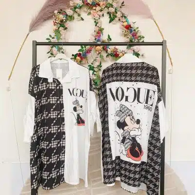 Zwei Lip Longtunika Kleider (Kopie) mit Minnie-Maus-Motiven hängen an einem Kleiderständer unter einem Blumenkranz.