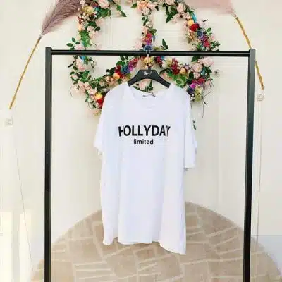 Weißes NJC-Hemd (Kopie) mit „Hollyday Limited“-Aufdruck, das an einem Regal vor einem Blumenkranz-Hintergrund hängt.