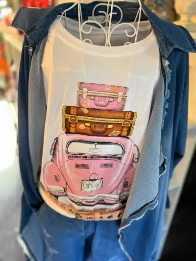 Ein Lets go-Shirt mit einem farbenfrohen Aufdruck von gestapeltem Gepäck und einem rosa Auto, das auf einem Kleiderbügel hängt und mit einer blauen Jeansjacke drapiert ist. Das Bild wurde in einer Einzelhandelsumgebung aufgenommen.