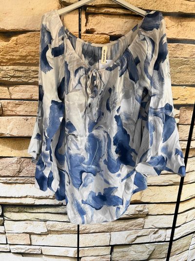 Blau-weiß gemusterte Graffic-Tunika-Bluse, die an einem Metallgestell an einer Steinwand hängt.