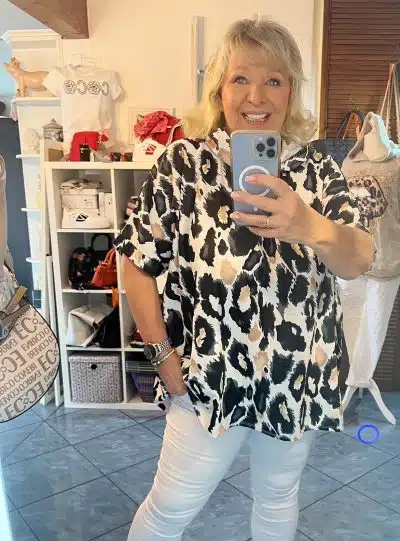 Eine lächelnde Frau, die in einer Boutique ein Selfie macht, mit einer Leo-Hoodie-Tunika im Hintergrund.