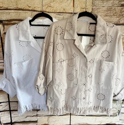 Zwei weiße Blusen im Lochstickerei-Stil hängen nebeneinander vor einem Steinmauerhintergrund, ergänzt durch eine Hole Summer Jeans Jacke.