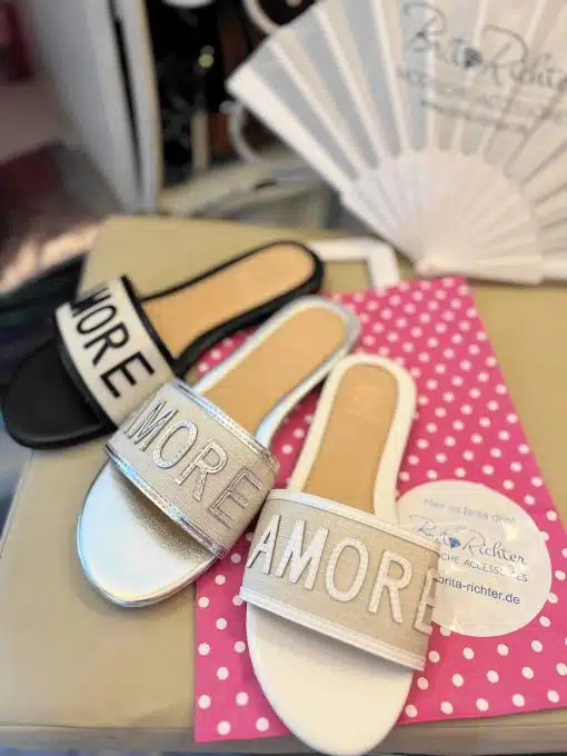Satz mit Produktname: Ein Paar modische Gucci-Turnschuhe mit dem Wort „Gucci“ auf einer gepunkteten Oberfläche, neben ähnlich gestalteten Sandalen mit Text und Werbematerialien im Hintergrund.