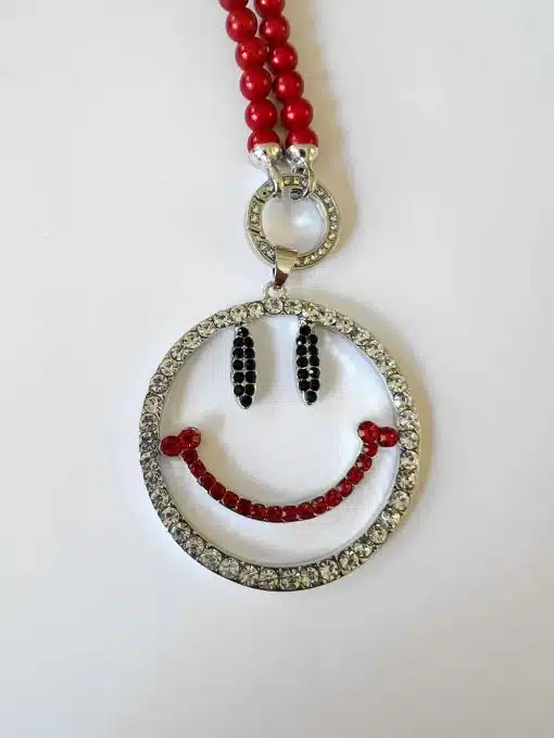 Ein Anhänger im „Hole big Smile Wechselanhänger“-Design, verziert mit roten Perlen, schwarzen Akzenten und klaren Kristallen an einer roten Perlenkette vor weißem Hintergrund.