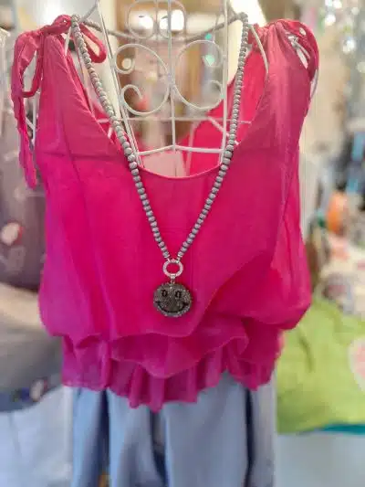 Ein rosa Seiden-Top mit Rüschendetails am unteren Ende hängt auf einem Kleiderbügel und wird durch eine türkisfarbene Perlenkette mit einem runden Anhänger akzentuiert.