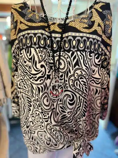 Eine Nahaufnahme einer Memory-Tunika mit komplizierten Mustern, die auf einem Kleiderbügel hängt und mit einer Perlenkette geschmückt ist.