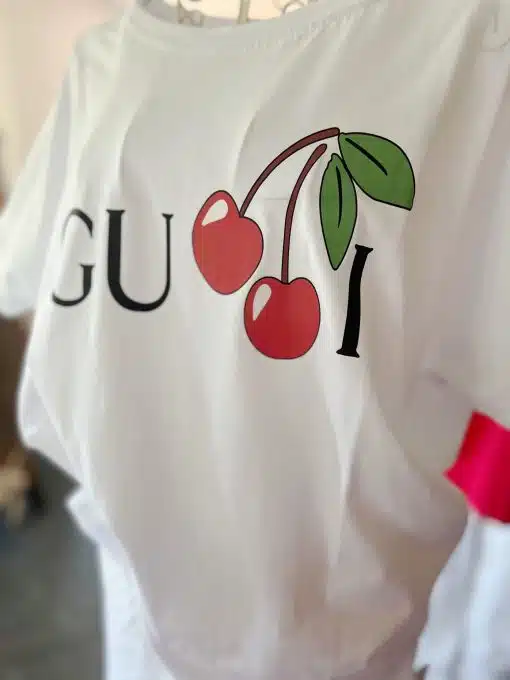 Weißes Kirsch-Shirt mit einer verspielten Variante eines Designer-Logos, bei dem das Wort „Gucci“ so abgeändert wurde, dass es ein Paar roter Kirschen als Buchstaben „C“ enthält.