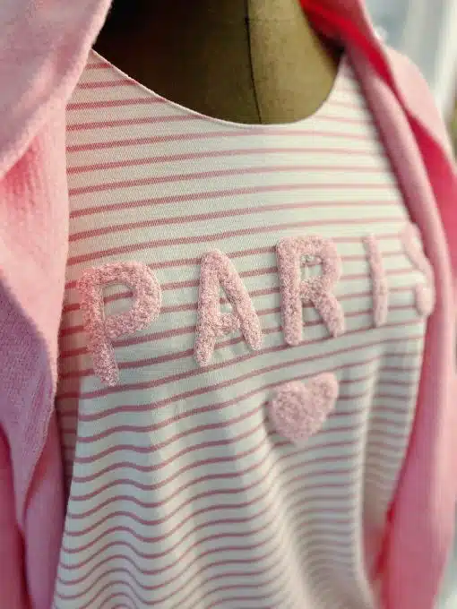 Nahaufnahme einer Schaufensterpuppe, die das Paris Stripe Shirt mit dem Wort „PARIS“ und einem Herzsymbol in strukturierten Buchstaben trägt. Ein passender rosa Kapuzenpullover liegt über ihren Schultern und vervollständigt den stylischen Look.