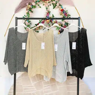 Vier Pullover hängen an einem Kleiderständer, in den Farben von links nach rechts: anthrazitfarbener Cherry Lurex Pulli, Beige, Hellgrau und Schwarz, jeweils mit Preisschildern. Ein Blumenkranz