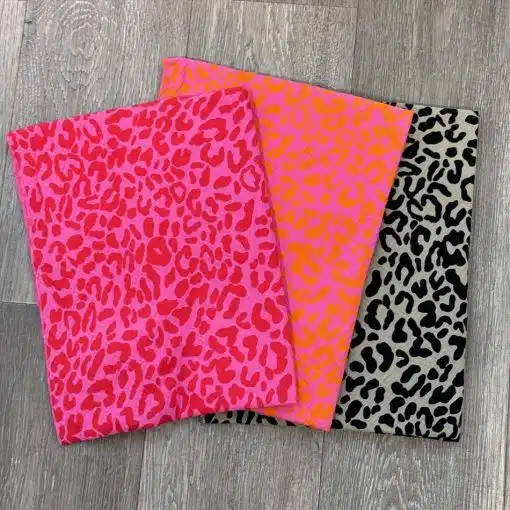 Satz mit Produktname: Drei Stücke von Sabrina Tuch mit Leopardenmuster, eines in leuchtendem Pink, ein anderes in fluoreszierendem Orange und das dritte in Schwarz und Grau, alle auf einer hellgrauen Holzoberfläche präsentiert.