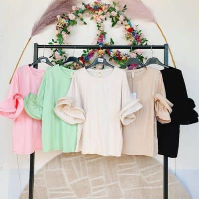 Vier Cherry´s Shirts in den Pastelltönen Rosa, Grün, Creme und Schwarz hängen auf Kleiderbügeln unter einer Blumenkranzdekoration vor einem weißen Hintergrund.