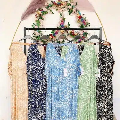 Eine Kollektion farbenfroher Tunika-XL-Kleider im italienischen Stil hängt an einem Ständer, darüber ein dekorativer Blumenkranz und eine große Feder. Die Szenerie scheint drinnen zu sein, möglicherweise in einer Boutique.