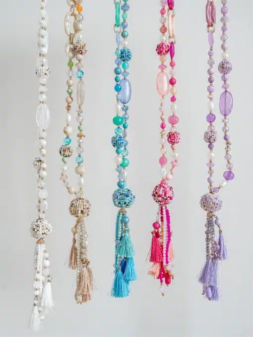 Eine Kollektion farbenfroher Perlenketten vom Typ „Ball Boho Kette“ mit vertikal hängenden Quastenenden vor einem weißen Hintergrund.