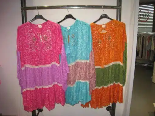 Drei farbenfrohe Kleider vom Typ Suntime Boho Kleid XL hängen in einem Geschäft an Ständern; rosa, türkis und orange, jedes mit detaillierten Mustern und gewellten Säumen.