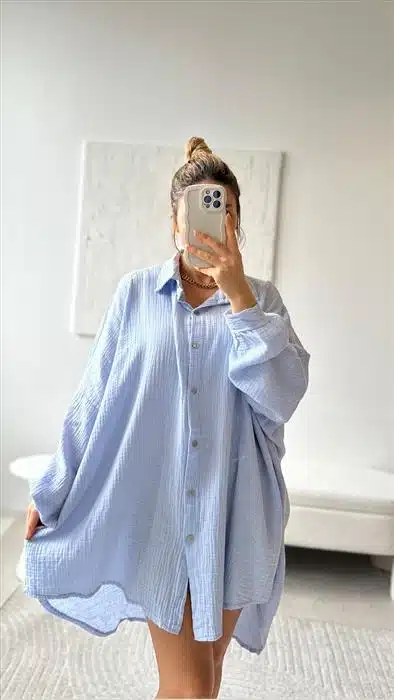 Eine Person macht ein Spiegel-Selfie, während sie eine große blau-weiß gestreifte Musselin-Bluse trägt. Die Kulisse ist ein moderner Raum mit einer schlichten weißen Wand und einer minimalistischen Bank.