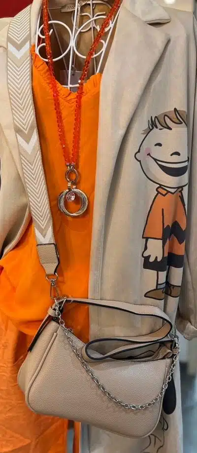 Eine Schaufensterpuppe trägt einen schicken beigen Trenchcoat mit Cartoon-Motiv über einem orangefarbenen Kleid. Als Accessoires trägt sie eine orangefarbene Halskette, einen Ringanhänger und eine silberne City Bag.