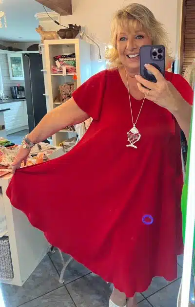 Eine Frau in einem roten Kleid von Miss Chiara Kleid macht ein Selfie in einem Spiegel. Sie lächelt und streckt den Arm aus, um das Foto aufzunehmen. Sie scheint in einem Brunnen zu sein.