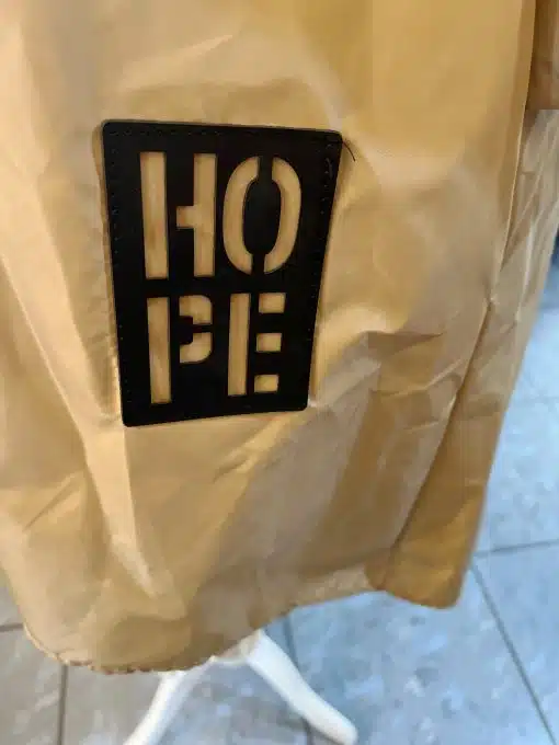 An einer beigen Hope Allwetterjacke ist ein schwarzes Badge mit der Aufschrift „Hope“ in weißen Blockbuchstaben befestigt.