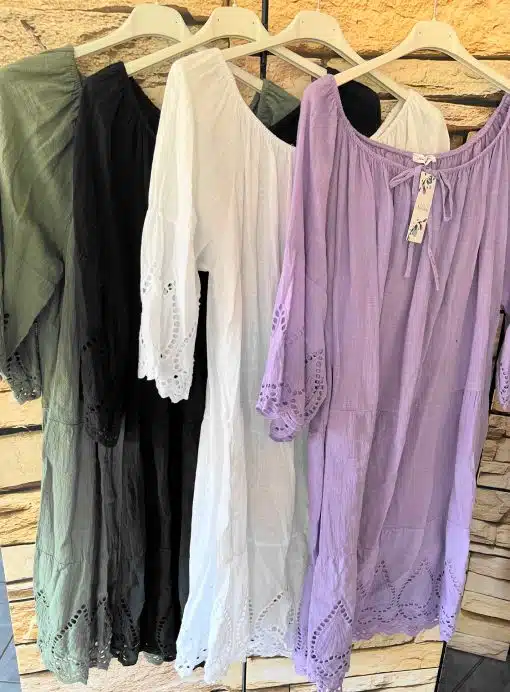 Eine Auswahl an Boho-Kleidern XL uni mit Spitzendetails, präsentiert auf Kleiderbügeln vor einer strukturierten Wand, in den Farben von links nach rechts: Olivgrün, Weiß und Lavendel.