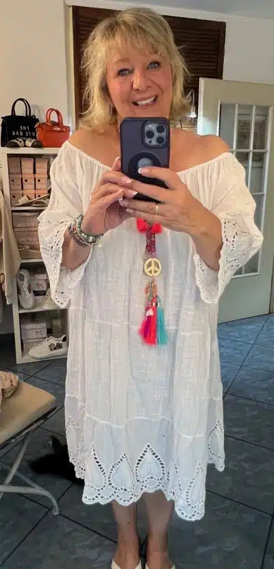 Eine Frau in einem weißen Boho Kleid XL Uni lächelt und macht mit ihrem Handy ein Selfie vor einem Spiegel.