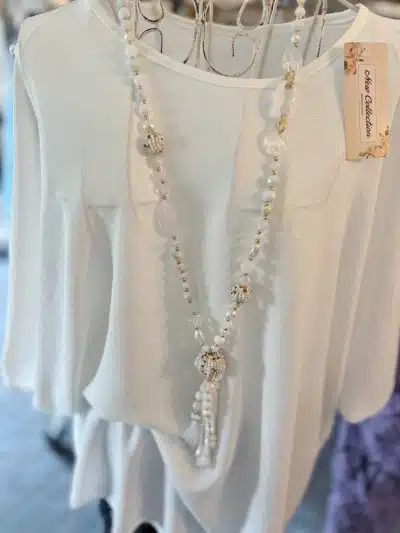 Eine weiße Bluse hängt auf einem Bügel und darüber hängt eine lange Ball Boho Kette mit filigranen Anhängern. Die Kulisse erinnert an eine Boutique mit verschwommenem Hintergrund.