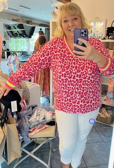Eine Frau in einem knallpink-roten „Leo Allover Neon“-Pullover und weißen Hosen macht in einer Boutique ein Selfie, umgeben von verschiedenen Kleidungsstücken und Accessoires.