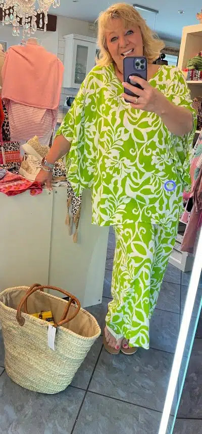 Eine Frau in einem leuchtend grün-weiß gemusterten Apple Blossom-Kleid macht in der Umkleidekabine einer Boutique ein Selfie und steht dabei neben einem geflochtenen Korb und einem Spiegel.