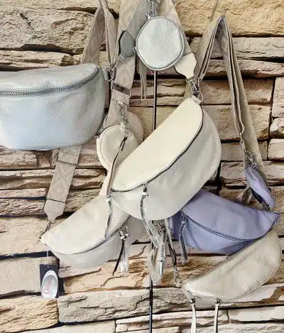 Eine Auswahl pastellfarbener Gürteltaschen, die an Haken vor einer Steinmauer hängen. Die Taschen sind in Beige-, Grau- und Lavendeltönen erhältlich und umfassen Prima Cross Over Taschen in verschiedenen Größen und Ausführungen.