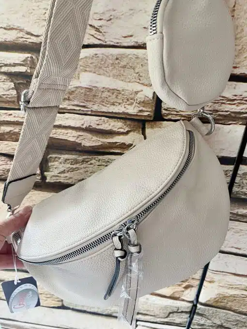 Eine Umhängetasche aus weißem Leder mit strukturierter Oberfläche hängt an einem Metallständer vor einer Backsteinwand. Die Tasche verfügt über einen verstellbaren Riemen und einen auffälligen silbernen Reißverschluss. Diese Prima Cross Over Tasche ist beides