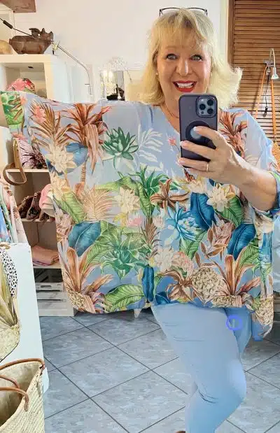 Eine Frau mit blondem Haar lächelt und macht ein Selfie in einem Spiegel. Sie trägt hellblaue Hosen und ein farbenfrohes Sommerkleid mit Blumenmuster und hält ein Smartphone in der Hand. Ein sonnendurchfluteter, mit Dekor gefüllter Raum.