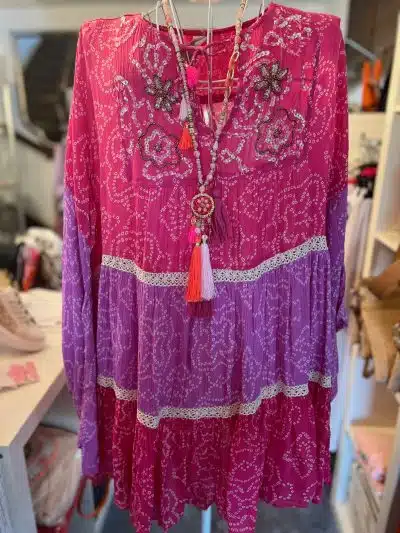 Ein farbenfrohes Suntime Boho Kleid XL mit detaillierter Stickerei und einer daran hängenden Quastenkette. Das Kleid hat leuchtend rosa und lila Lagen, die mit Spitze und Blumenmustern verziert sind.