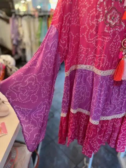 Ein leuchtend rosa Kleid mit detailliertem weißen Paisleymuster und Spitzenbesatz (Suntime Boho Kleid XL) hängt in einem Bekleidungsgeschäft. An der Taille ist eine Quaste befestigt, im Hintergrund sind andere Kleidungsstücke unscharf.