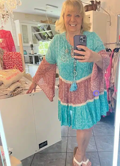 Eine Frau mit blondem Haar macht in der Umkleidekabine einer Boutique ein Selfie. Sie trägt ein türkis-rosa Suntime Boho Kleid XL und weiße Sandalen und lächelt in die Kamera ihres Telefons.