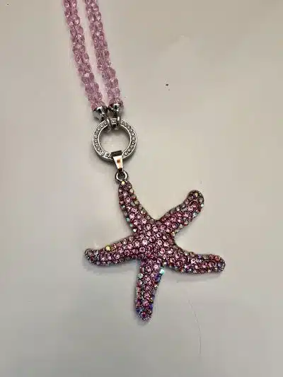 An einer rosa Perlenkette hängt vor einem blassen Hintergrund ein rosa und lila Seestern-Anhänger mit Juwelen.