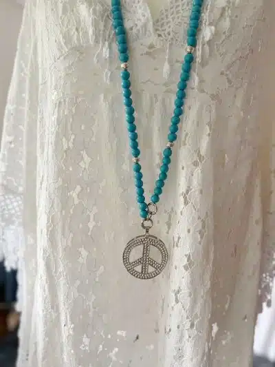 Eine türkisfarbene Perlenkette mit einem silbernen Friedenssymbol-Anhänger hängt an einem Beachtime-Poncho, der in einem Raum mit Tageslicht ausgestellt ist.