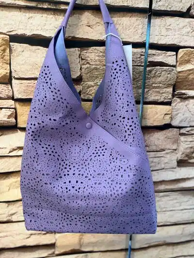 Eine lilafarbene Modetasche mit aufwendigen lasergeschnittenen Mustern hängt an einer Backsteinwand. Die Tasche verfügt über einen Knopfverschluss und einen Schlaufengriff.