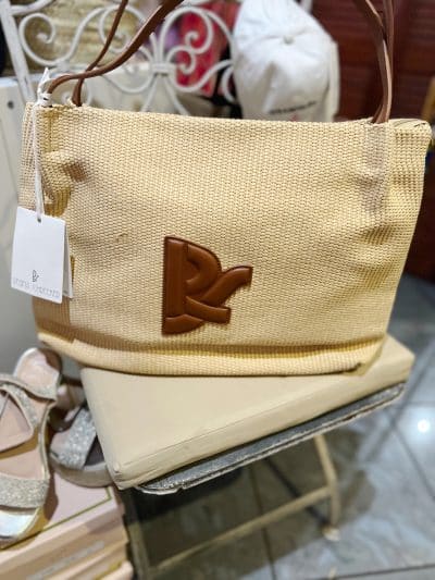 Eine beige Regina Schrecker Tasche mit einem auffälligen braunen Buchstaben „R“ auf der Vorderseite, ausgestellt auf einem Glastisch in einem Laden. Die Handtasche hat eine strukturierte Oberfläche und.