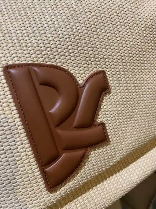 Nahaufnahme einer braunen „p“-Applikation aus Leder, die auf einem gewebten beigen Hintergrund einer Regina Schrecker Tasche befestigt ist und detaillierte Nähte und Textur zeigt.