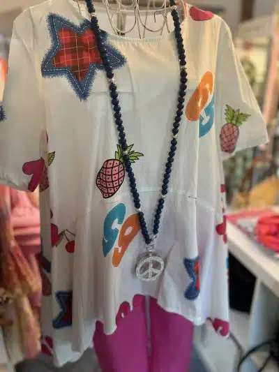 Ein langes weißes Hemd aus italienischer Baumwolle mit farbenfrohem Peace-Zeichen und Ananas-Aufdruck, präsentiert auf einer Schaufensterpuppe, kombiniert mit leuchtend rosa Hosen und als Accessoire eine blaue Perlenkette mit einem großen Peace-Symbol.