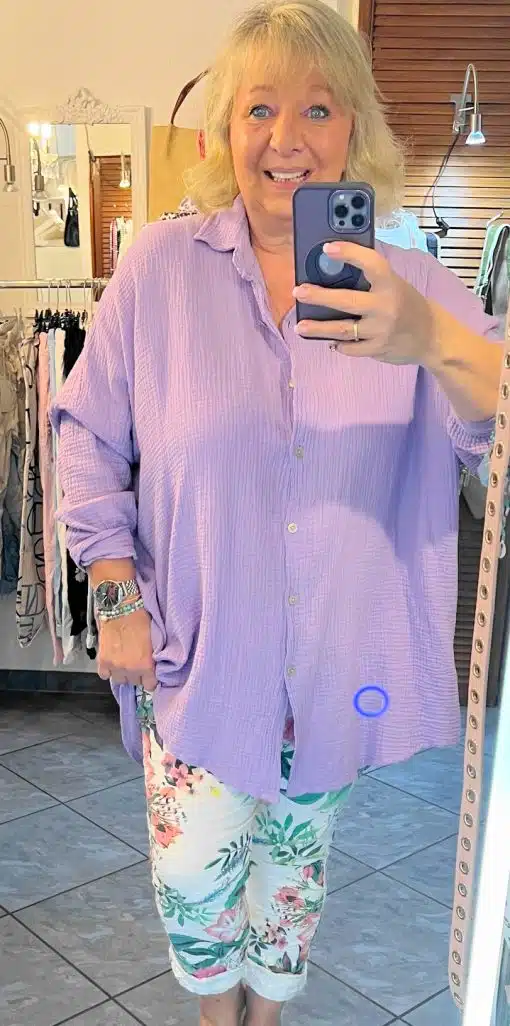Satz mit Produktnamen: Eine Frau mit blondem Haar macht ein Selfie vor einem Spiegel. Sie trägt eine lila, übergroße Musselin-Bluse und geblümte Hosen und lächelt in einem hell erleuchteten Raum mit Kleiderständern im Hintergrund.