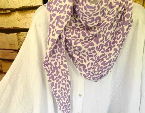 Weißes Hemd auf einem Bügel, geschmückt mit einem violett-weißen Dreiecktuch aus Musselin mit Tiermuster, das um den Halsausschnitt drapiert ist. Der Hintergrund besteht aus Steinziegeln.