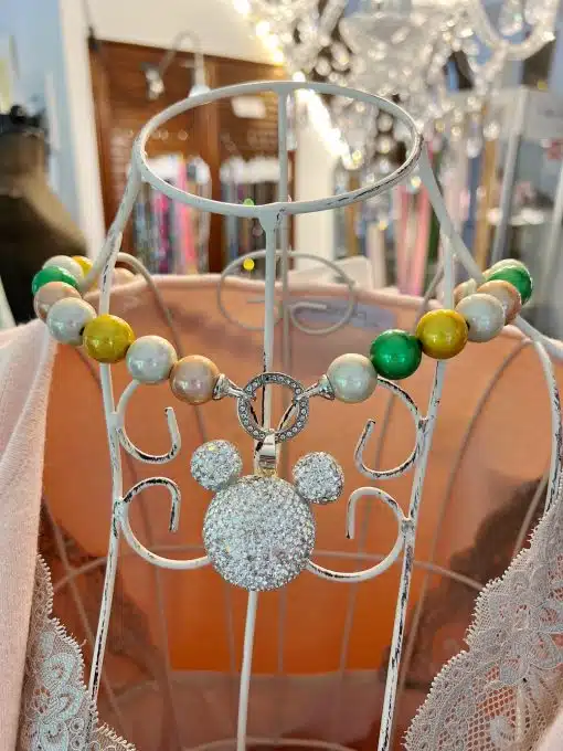 Eine magische Wechselkette kurz mit großen Perlen in Grün, Gelb und Perlmutt wird auf einer weißen Metallpuppe präsentiert. Die Kette hat einen großen, funkelnden Mickey-Mouse-Kopfanhänger. Im Hintergrund ist ein sanft beleuchteter Laden mit verschiedenen Artikeln zu sehen.