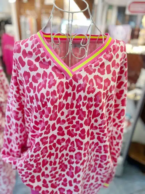 Ein langärmliger „Leo Allover Neon“-Pullover mit rosa-rotem Leopardenmuster hängt auf einem Drahtbügel. Der V-Ausschnitt ist mit rosa-gelben Streifen eingefasst, während im Hintergrund verschwommene Artikel zu sehen sind.