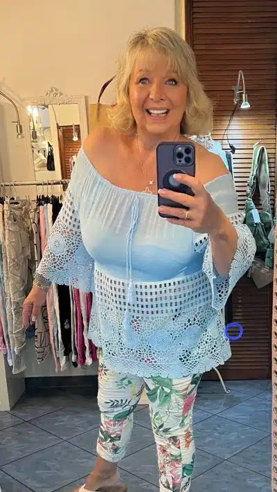 Eine Frau mit blondem Haar lächelt, während sie in einer Boutique ein Selfie macht. Sie trägt eine hellblaue Welcome-Tunika (Kopie) und weiße Hosen mit Blumenmuster.
